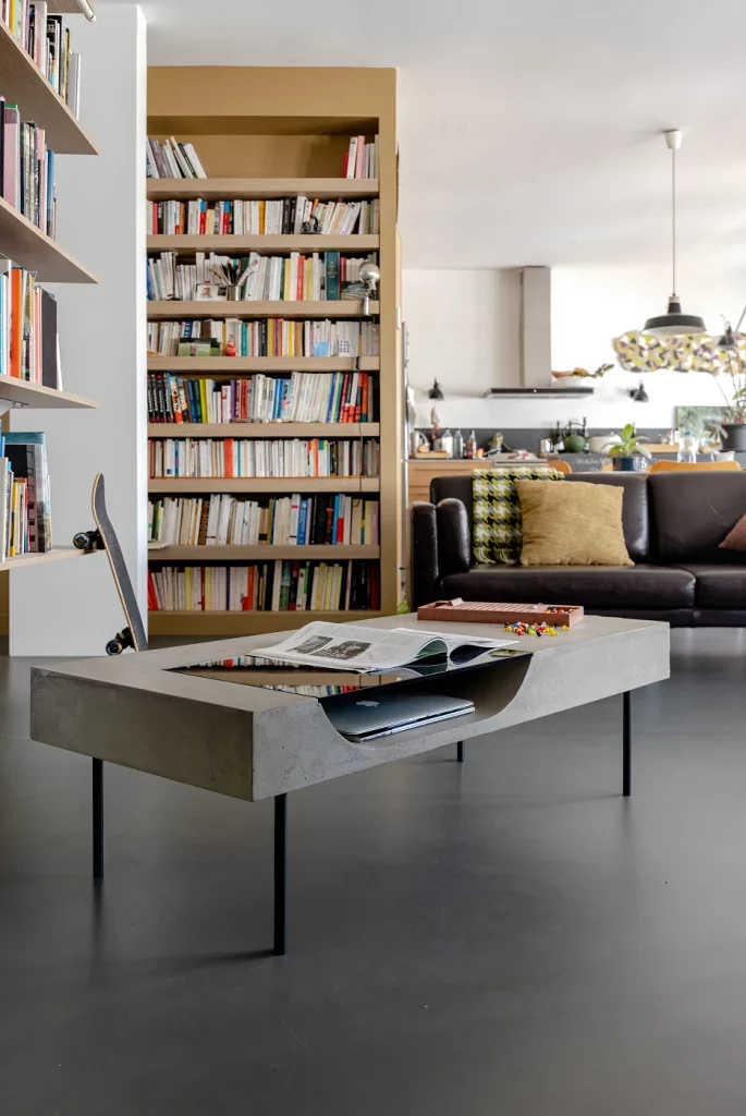 Table basse Curb en béton avec une niche en verre, dans un salon avec une bibliothèque.