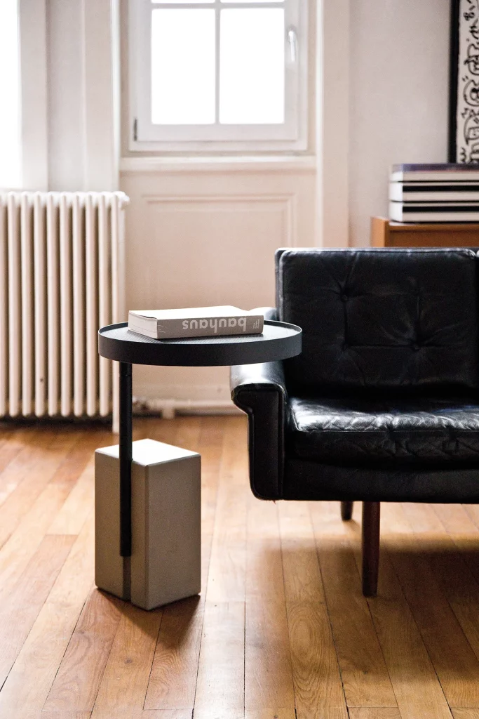 Table d’appoint Twist avec un plateau en métal noir sur une base en béton, à côté d’un fauteuil en cuir.