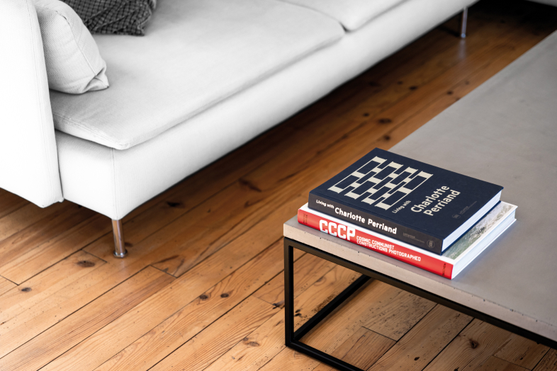 La table basse minimaliste en béton et acier noir devant un canapé blanc modern surmonté d'un livre sur Charlotte Perriand